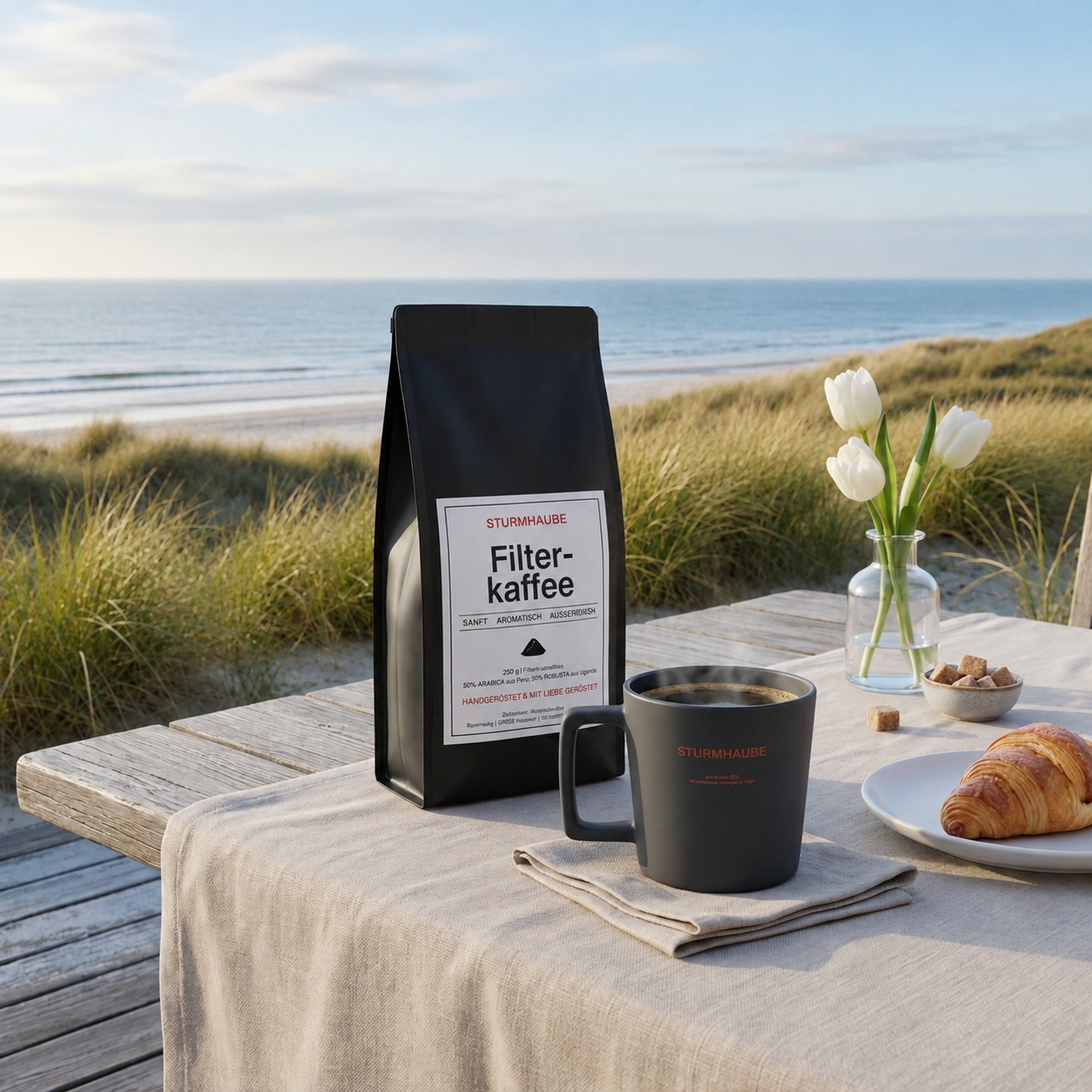Schwarze Packung STURMHAUBE Filterkaffee und schwarze Kaffeetasse auf einem rustikalen Holztisch mit Croissant und weißen Tulpen. Im Hintergrund sorgen sommerlich grüne Dünen und der Blick auf das Meer für eine entspannte Küstenatmosphäre.