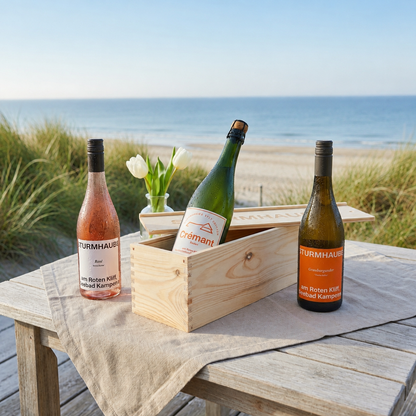 Eine geöffnete Holz-Weinkiste mit einer Flasche Sturmhaube Crémant sowie einer Rosé- und einer Grauburgunderflasche steht auf einem Holztisch direkt am Strand von Sylt. Im sommerlichen Hintergrund sind grüne Dünengräser, das Meer und weiße Tulpen zu sehen. Die Szene vermittelt eine entspannte Küstenatmosphäre und eignet sich als hochwertiges Wein-Geschenkset.