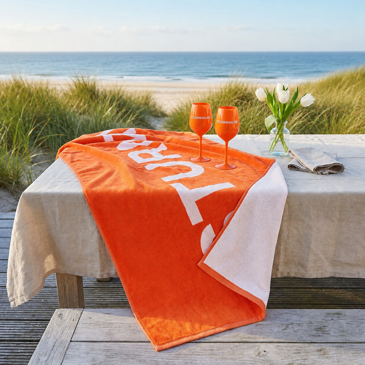 Ein orangefarbenes Sturmhaube-Strandtuch liegt dekorativ auf einem mit Leinentuch gedeckten Holztisch am Strand von Sylt. Daneben stehen zwei orangefarbene Sturmhaube-Weingläser sowie eine Vase mit weißen Tulpen. Im sommerlichen Hintergrund sind grüne Dünengräser, der Sandstrand und die Nordsee zu sehen.